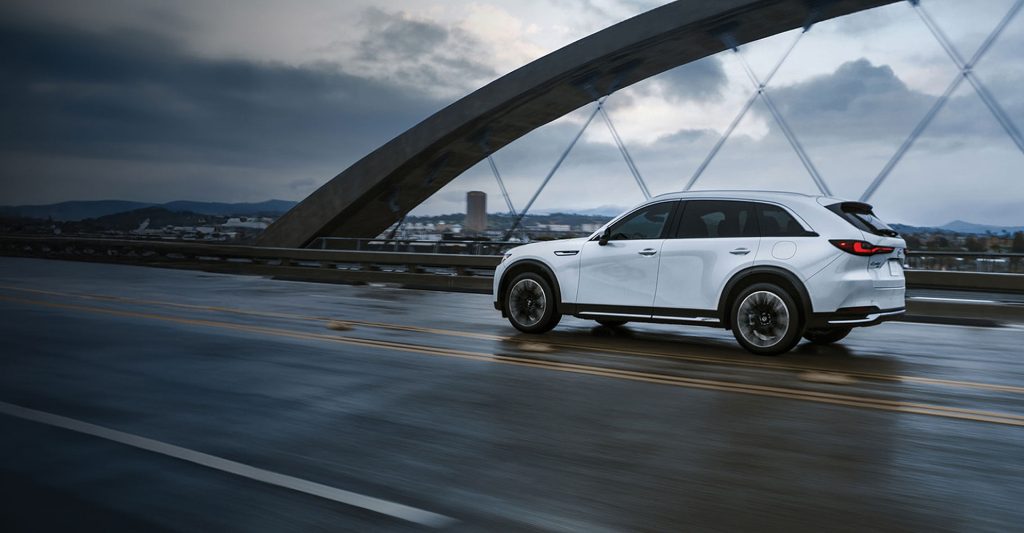 A white Mazda CX-90 drives across a wet bridge under a cloudy sky.