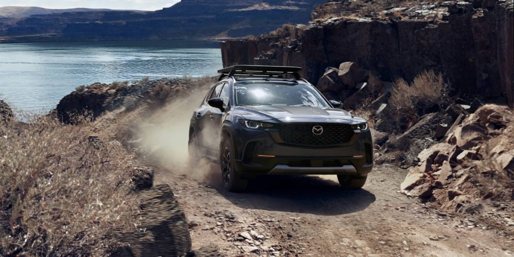 A dark SUV kicks up dust while driving along a rocky lakeside trail.