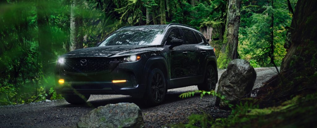 A black SUV drives along a wet forest road surrounded by dense green trees and moss-covered rocks.