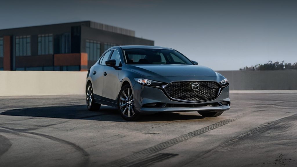Front three-quarter view of a dark gray Mazda3 Sedan parked on an outdoor concrete rooftop lot with a modern dark building structure in the background.