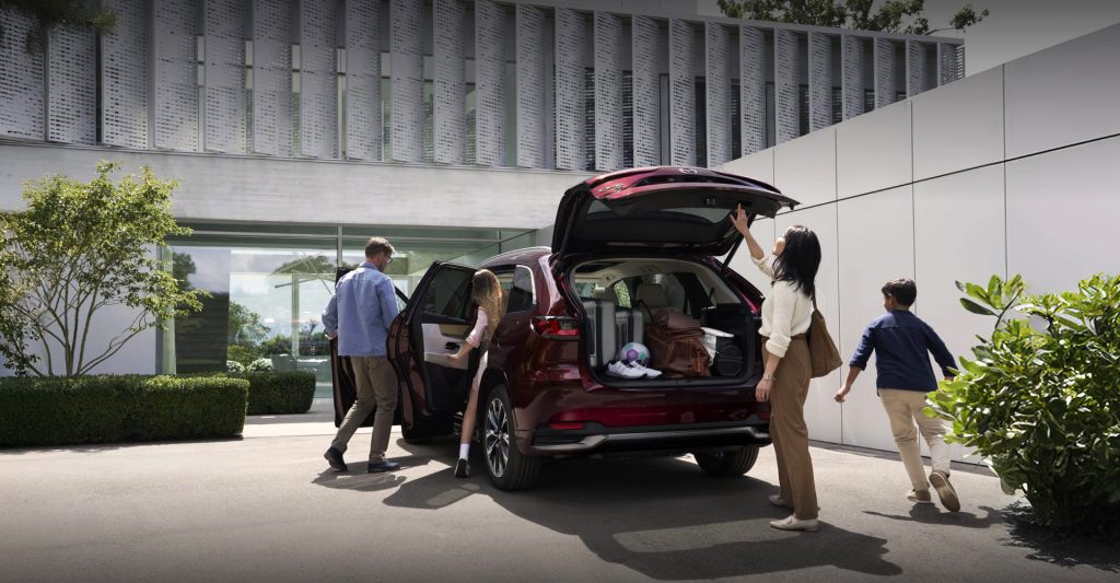 Family loading luggage into a 2026 Mazda CX-90 SUV parked outside a modern home, showcasing spacious cargo capacity and everyday versatility.